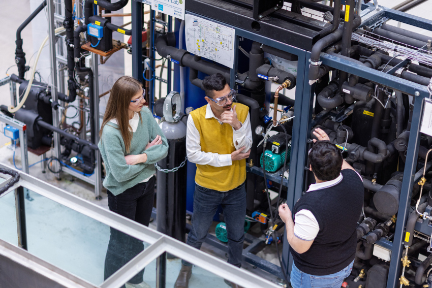 Tre studenter står ved en stor varmepumpe ved varmetekniske laboratorier