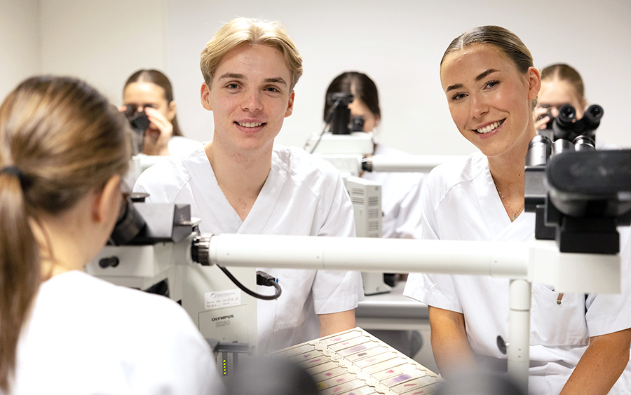 Two students smiling in a laboratory with microscopes and other students