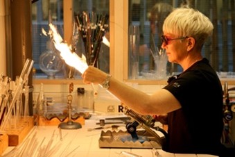 A glassblower sits at a table and works with glass and flame.
