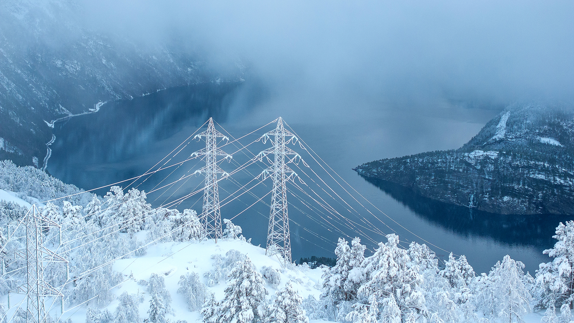  High voltage power lines over a fjord in a mountainous terrain with snow.