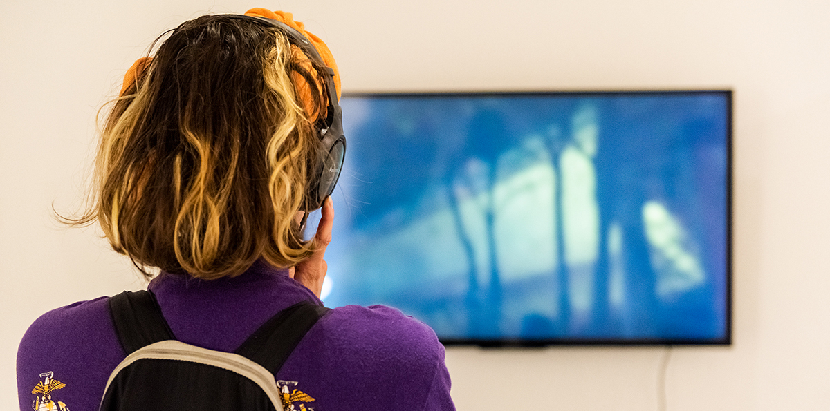 A photo of a woman from behind. She is wearing headphones and is looking at an art video.